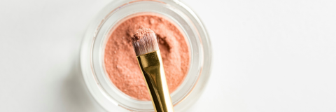 A glass ramekin on white background with orange powder inside it with a brush soaked into it